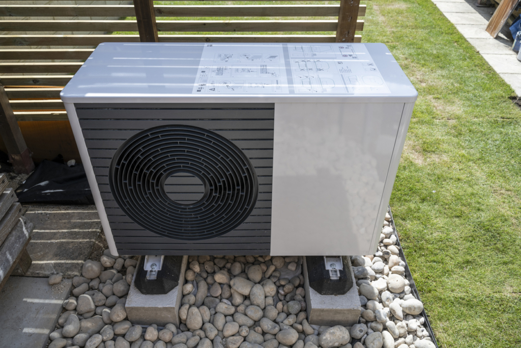 An outdoor air source heat pump unit is installed on concrete supports and surrounded by pebbles in a garden area, with signs of careful landscaping such as a wooden fence and green grass visible in the background.