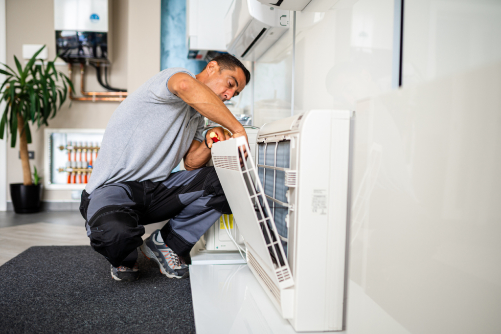 A technician in work clothes kneels indoors while servicing a wall-mounted HVAC unit, using a screwdriver on the open front panel. A plant and equipment can be seen in the background.