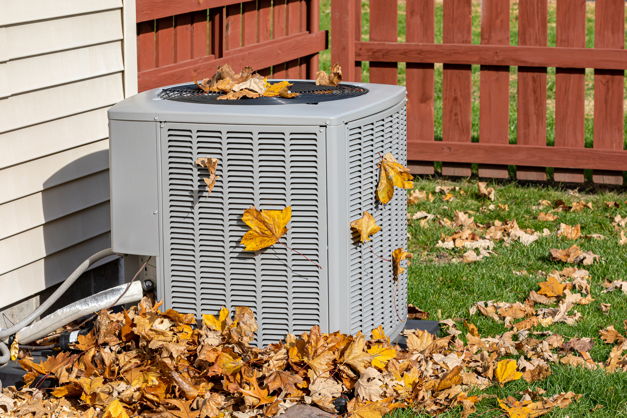 A gray outdoor air conditioning unit in a Midwest yard is covered with fallen autumn leaves; more leaves are piled around its base beside a beige house and red wooden fence, with green grass nearby.