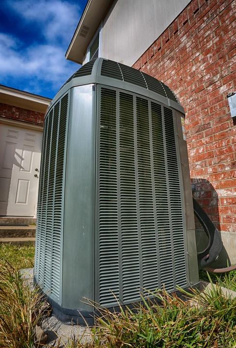 A large outdoor air conditioning unit sits next to a brick house, surrounded by grass, with a clear blue sky overhead.