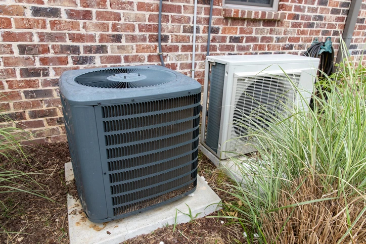 Two outdoor air conditioning units sit on concrete pads beside a brick house, surrounded by plants and grass. One unit is gray and the other is white.