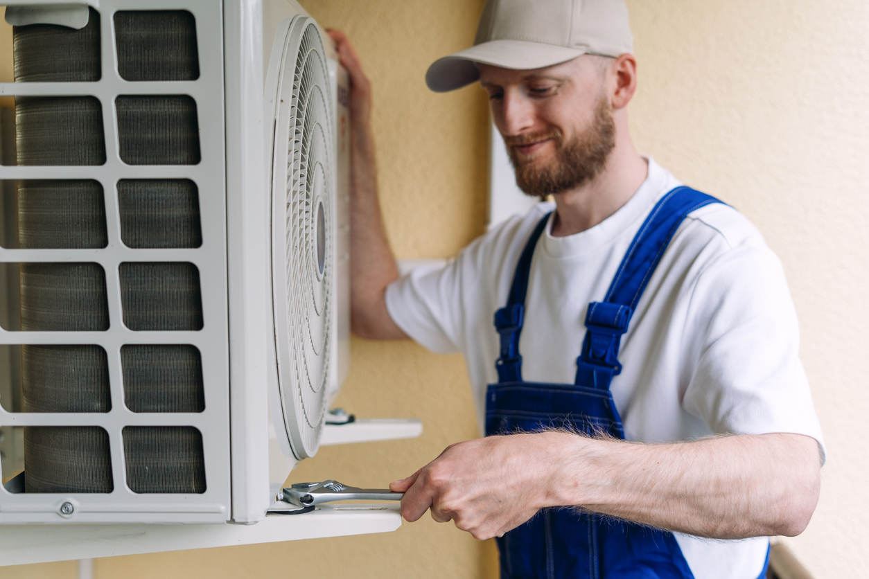 A man wearing blue overalls and a beige cap uses a wrench to work on an outdoor air conditioning unit mounted on a wall.