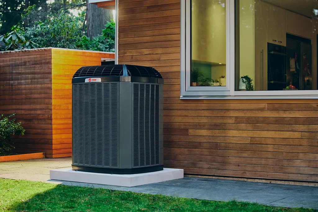 A large air conditioning unit is installed on a concrete pad next to a modern wooden house, beneath a window, with green grass and trees visible nearby.