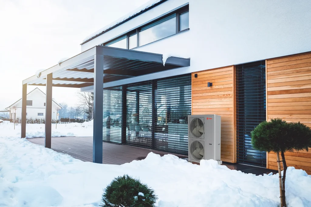 A modern house with wood and glass exterior sits in a snowy landscape. A covered patio with outdoor furniture is visible, as well as a heat pump unit mounted on the wall near small shrubs.