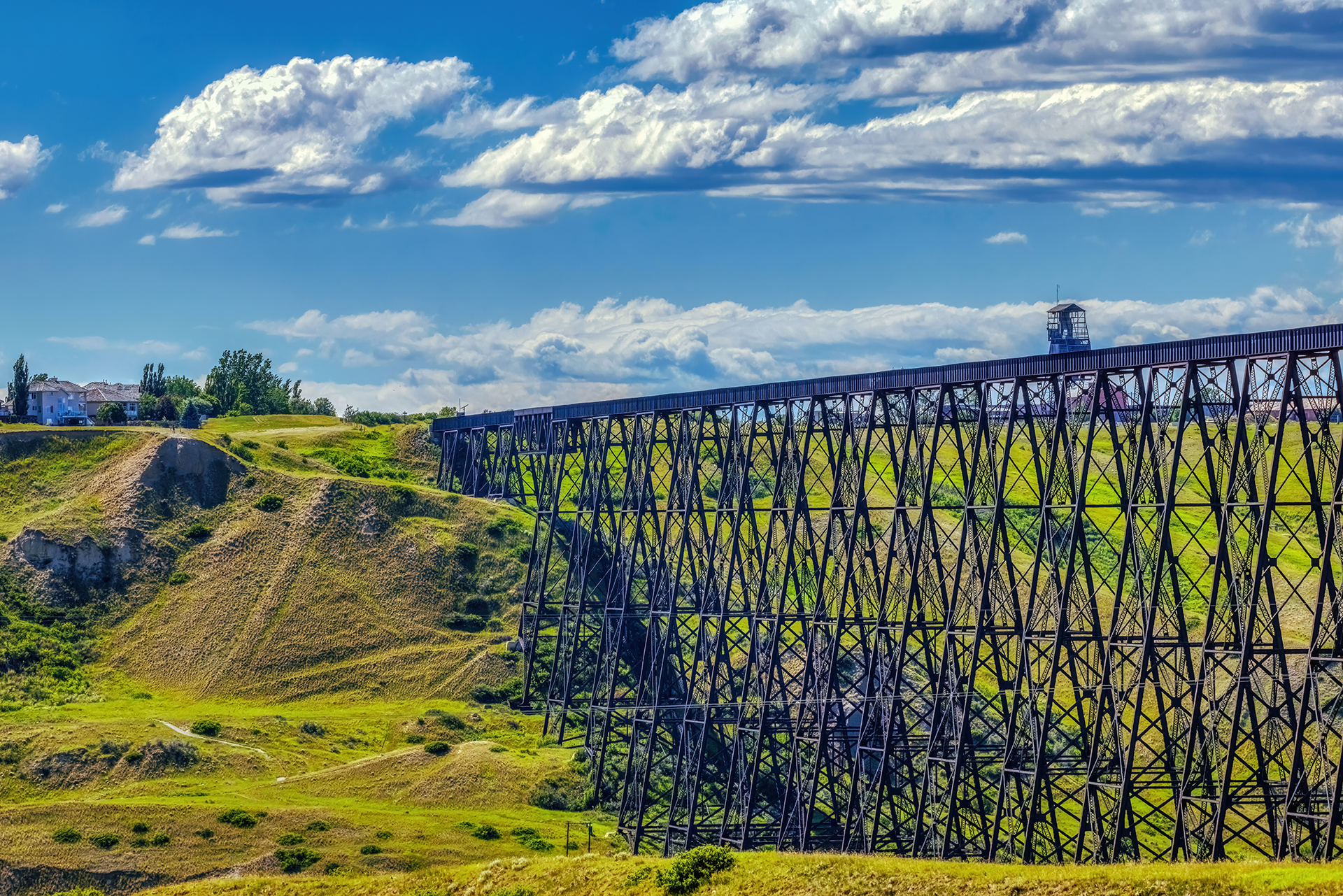 A tall steel railway trestle bridge crosses a grassy, hilly landscape under a partly cloudy blue sky, with trees and buildings visible in the distance.