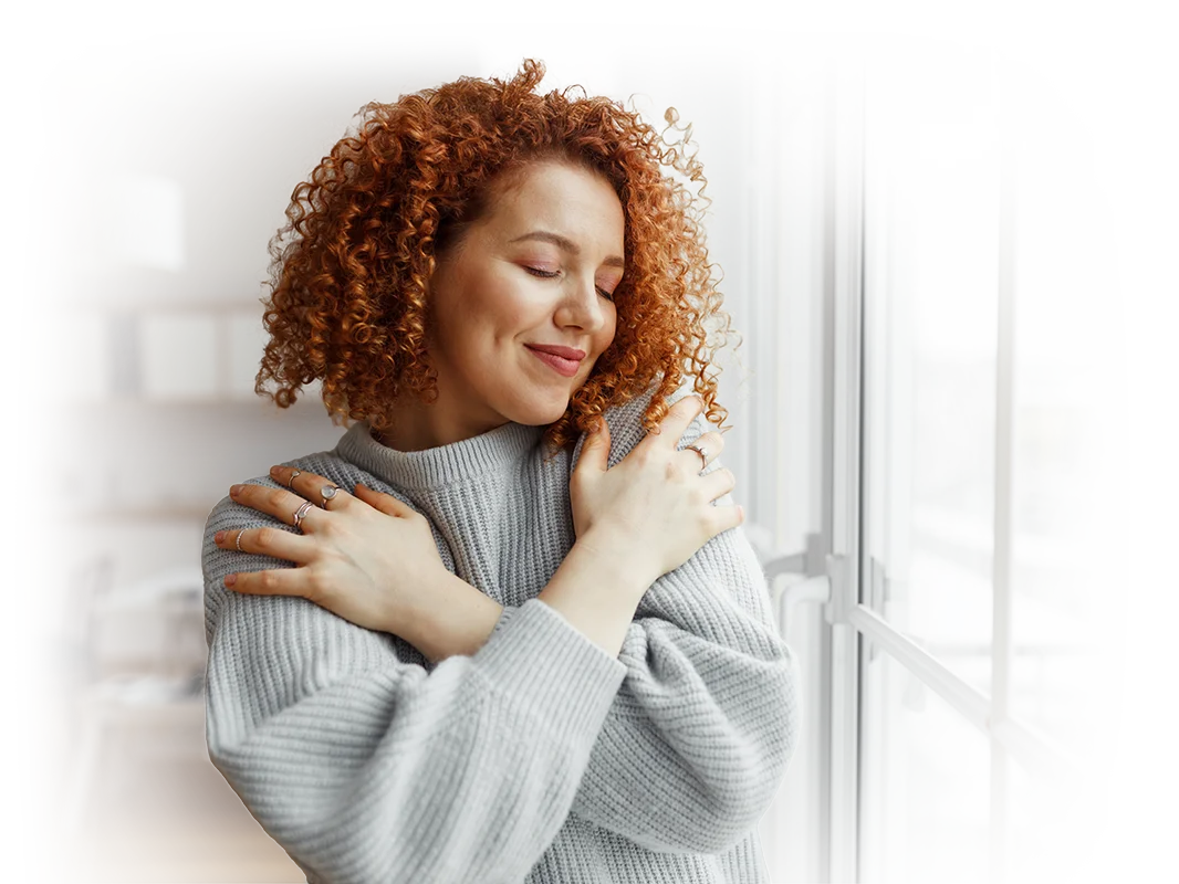 A woman with curly red hair and a gray sweater stands by a window, eyes closed and smiling gently, hugging herself in a cozy, sunlit room.