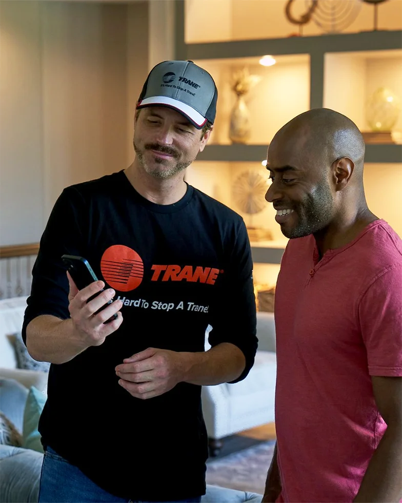 Two men are standing indoors, smiling and looking at a smartphone. One man is wearing a black Trane shirt and a Trane cap, while the other is wearing a red t-shirt. Shelves and decor are visible in the background.