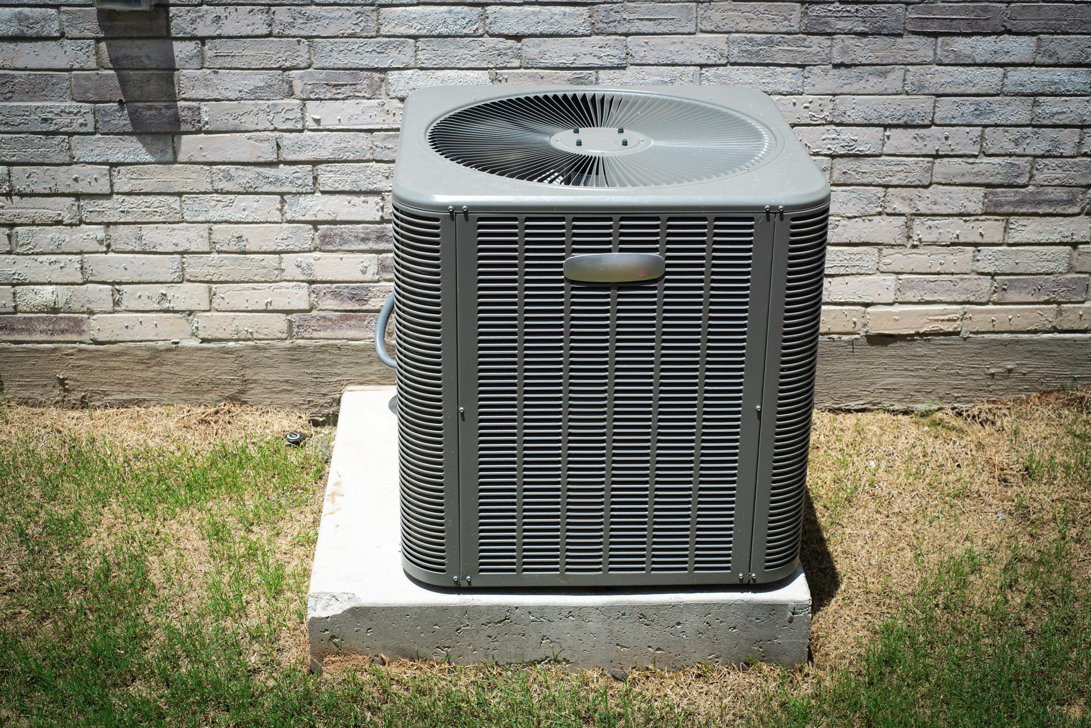 A gray outdoor air conditioning unit sits on a concrete pad next to a brick wall, surrounded by grass.