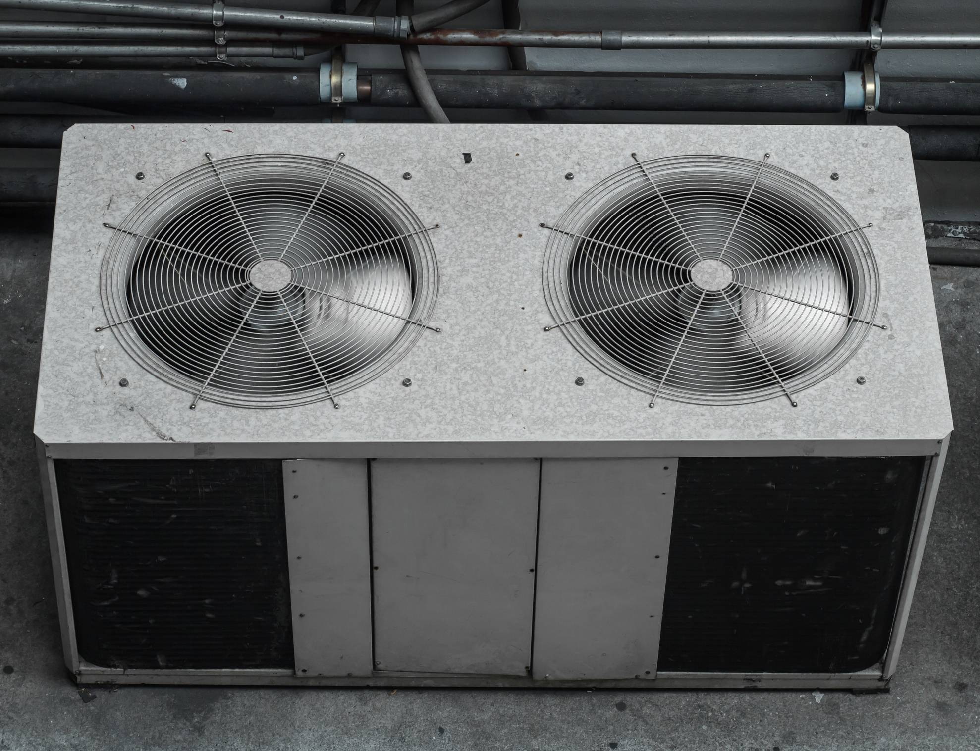 A top view of a large industrial air conditioning unit with two metal fans, installed outdoors on a concrete surface, with pipes and cables visible in the background.